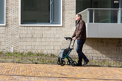 An individual walks outside using a rollator, near a brick building with large windows.